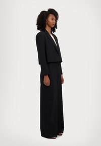 Blazer noir cropped avec un bouton, associé à un pantalon large. Tissu lisse et design structuré. Le modèle se tient de profil.