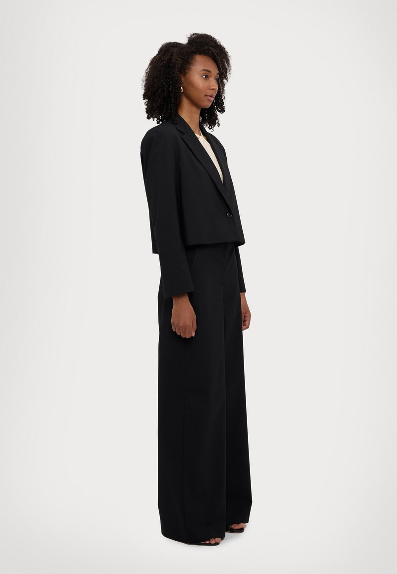 Blazer noir cropped avec un bouton, associé à un pantalon large. Tissu lisse et design structuré. Le modèle se tient de profil.