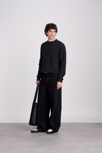 Zwarte gebreide trui, losse zwarte broek en zwarte tas. Sneakers met witte zolen en donkere accenten bij de neus. Gladde texturen, minimalistisch ontwerp.