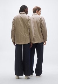 Két személy áll egymás háttal, bézs színű, kockás mintájú zakót és széles szárú sötét farmernadrágot viselnek, fehér tornacipővel.