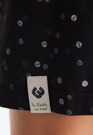 Czarna tkanina w kolorowe kropki z białą metką z napisem "W Ziemię wierzymy" pod okrągłym logo, blisko nogi osoby.