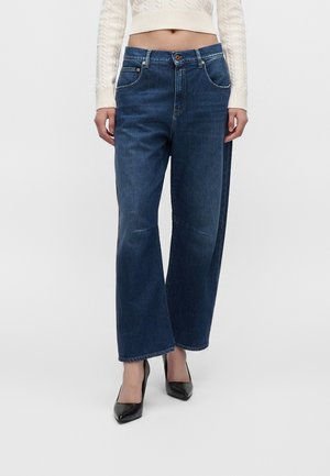 Frau trägt dunkelblaue Jeans mit weitem Bein, einen kurzen weißen Zopfmuster-Pullover und schwarze spitze High Heels vor einfarbigem Hintergrund.