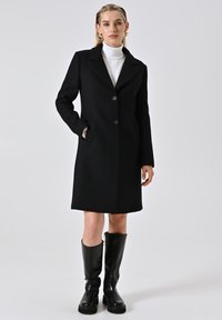 Cappotto di lana nero con colletto a revers, design a un bottone e due tasche anteriori. Modello abbinato a un dolcevita bianco e stivali neri fino al ginocchio.