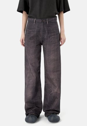 Pantalon à jambes larges avec motif à carreaux noirs et gris, porté avec des chaussures noires et une chemise noire à manches courtes, debout devant un fond uni.