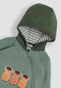 Kapuzensweatshirt in Olivgrün mit gestreiftem Innenfutter. Drei gestickte Cartoon-Kühe in Orange und Braun auf der Vorderseite. Mit Reißverschluss.