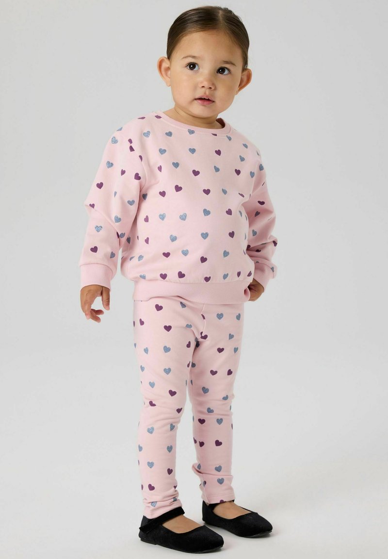 Completo con felpa rosa e leggings con piccoli motivi a cuore viola e blu, realizzato in tessuto morbido. Scarpe basse nere completano l'outfit.