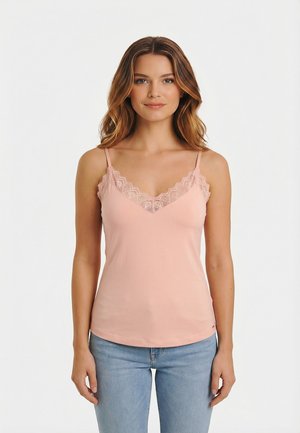 Giovane donna con capelli mossi che indossa una canottiera rosa con bordi in pizzo e jeans azzurri, in piedi su uno sfondo bianco semplice.