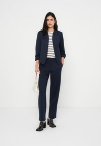 Ναυτικό μπλέ σακάκι και παντελόνι από λείο ύφασμα, σετ με ριγέ τοπ. Τα αξεσουάρ περιλαμβάνουν μια μικρή λευκή τσάντα και μαύρα παπούτσια με χοντρό τακούνι.