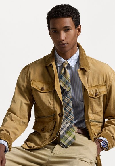 Καφέ βαμβακερό μπουφάν με μπροστινές τσέπες, σε συνδυασμό με ριγέ μπλε πουκάμισο και πουκάμισο με πλάγιο σχέδιο με πράσινα στοιχεία. Οι μπεζ παντελόνες ολοκληρώνουν την εμφάνιση.