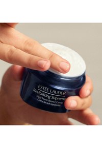Crème Revitalizing Supreme Night Power Bounce d'Estée Lauder dans un pot rond bleu foncé avec un couvercle blanc texturé, tenu entre deux mains.