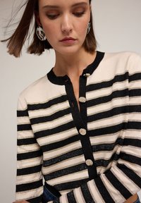 Svart och krämfärgad randig cardigan med ribbad struktur, metalliska detaljer, rund halsringning och fyra knappar framtill.