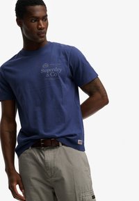 Joven mirando de lado con una camiseta azul de Superdry, pantalones cargo beige y un cinturón marrón sobre un fondo blanco liso.