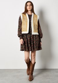 Brązowa haftowana kamizelka na tle czarnej sukienki w kwiaty z bufiastymi rękawami, w połączeniu z brązowymi kowbojkami. Płaskie tło, neutralne odcienie.