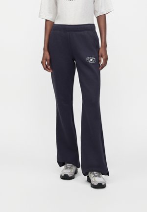 Persoon draagt marineblauwe sweatpants met wijde pijpen en New Balance-logo op de dij, gecombineerd met een wit shirt en grijze sneakers.