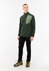 Man in een donkergroene zip-up jas met een lichtgroene borstpatch, zwarte broek en gele wandelschoenen, staande en kijkend naar rechts.