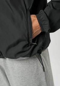 Käsi paigutatud musta jope taskusse, mis on kantud helehallide dressipükste peale, millel on nähtavad elastsed mansetid ja paelad.