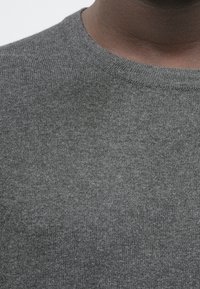 Κοντινό πλάνο ενός ατόμου που φοράει ένα γκρίζο πλεκτό πουλόβερ με λαιμόκοψη, που δείχνει τον λαιμό και την ανώτερη περιοχή του στήθους.