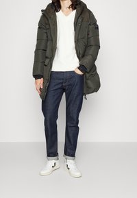 Nafouklá khaki parka s kapucí, přes bílý svetr s výstřihem do V, kombinovaná s tmavými džínami a bílými teniskami s černými detaily.