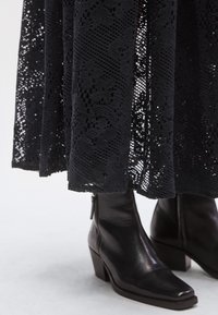 Μαύρα μποτάκια αστρα�γάλου με μυτερή μύτη, φτιαγμένα από λείο δέρμα, συνδυασμένα με μαύρο φόρεμα δαντέλας μέχρι το γόνατο με περίπλοκα σχέδια.