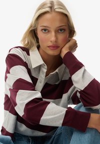 Blonde Frau mit blauen Augen, die ein weinrotes und graues gestreiftes Langarmshirt und Jeans trägt, und ihren Kopf auf ihre Hand stützt.