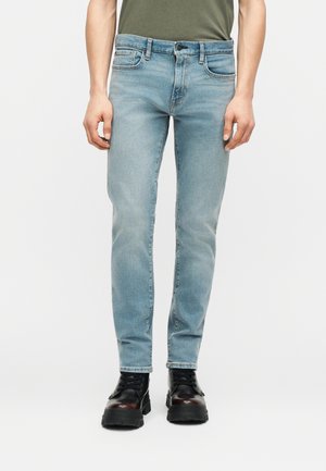 Muž v svetlomodrých úzkych rifle a čiernych členkových topánkach so šnúrkami stojaci pred bielym pozadím.