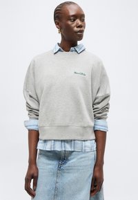 Γυναίκα με πλεξίδες στα μαλλιά, φοράει χρυσά σκουλαρίκια, ένα ανοιχτό μπλε ριγέ πουκάμισο κάτω από γκρι φούτερ Marc O'Polo και ανοιχτό μπλε τζιν παντελόνι.