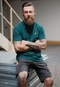 Camisa de polo de color verde azulado con mangas cortas y cuello abotonado, combinada con pantalones cortos gris oscuro que tienen un bolsillo lateral. Los tatuajes en los brazos son visibles.