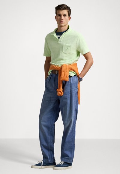 Lichtgroen polo shirt van gestructureerde stof, oranje gebreid sweater om de taille geknoopt, losse blauwe denim broek en marineblauwe sneakers.