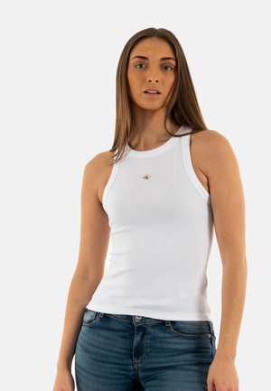 Top de tirantes blanco con textura acanalada, escote redondo y detalle de logo en el centro. Combinado con jeans de mezclilla azul, presenta un diseño ajustado y una textura suave.