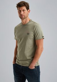 Olivegrünes Baumwoll-T-Shirt mit Rundhalsausschnitt, kurzen Ärmeln und einem Logodetail auf der Brust. Kombiniert mit dunklen Jeans.