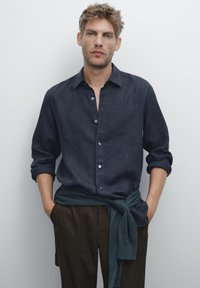 Man with tousled blond hair wearing a dark blue buttoned shirt, dark trousers, and a blue sweater tied around his waist.
