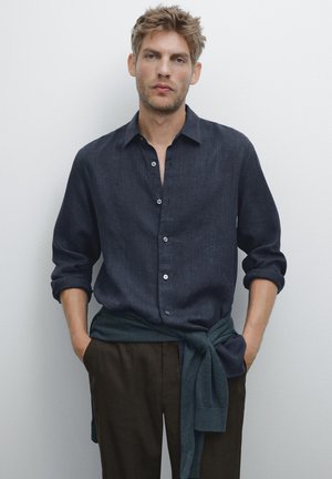 Man with tousled blond hair wearing a dark blue buttoned shirt, dark trousers, and a blue sweater tied around his waist.