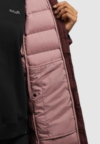 Veste matelassée bordeaux avec une doublure rose pâle, dotée d'une poche latérale et de sections cousues. Sweat-shirt noir en dessous.