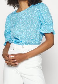 Top azzurro chiaro a maniche corte con motivo floreale bianco, maniche a puff arricciate e scollatura rotonda. Abbinato a jeans bianchi a vita alta.