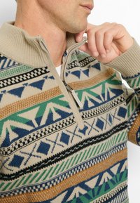 Beige Zip-Up-Pullover mit mehrfarbigen geometrischen Mustern in Blau, Grün, Orange und Schwarz. Verfügt über einen gerippten Kragen und strukturstrick.