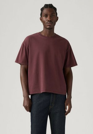 Mand med flettet hår iført en løs, kortærmet vinrød skjorte og mørkeblå jeans, stående mod en ensfarvet lys baggrund.