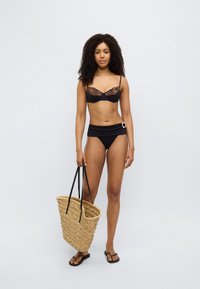 Μαύρο μπικίνι με δαντελένιες λεπτομέρειες, ψηλόμεσο κάτω μέρος και πλεκτή ψάθινη τσάντα με μαύρα λουριά. Το μοντέλο φοράει μαύρες σαγιονάρες.