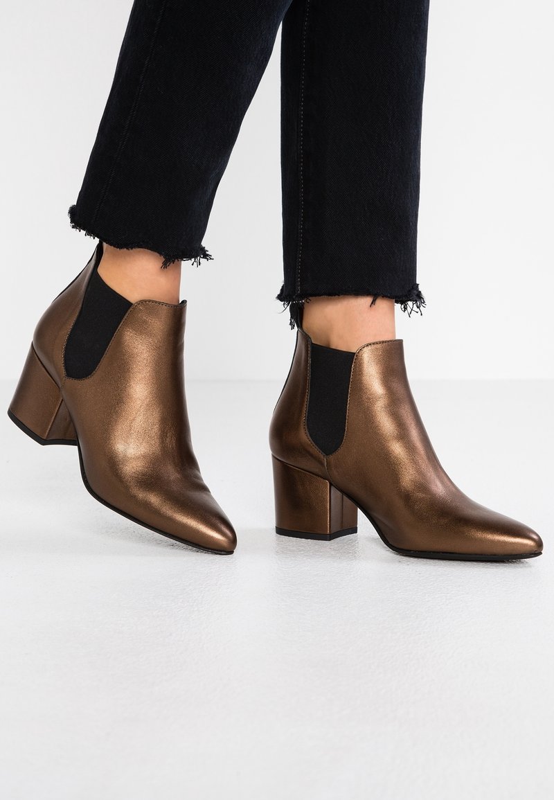 Femme portant des bottines métalliques bronze à talons blocs et panneaux latéraux élastiques noirs, associées à un jean noir court à bords effilochés.