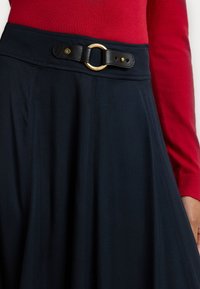 Mornarsko A-linijski krilo s črnim usnjenim pasom, ki ima zlato kovinsko zaponko; gladka tkanina z vidnimi šivi. Rdeča majica delno vidna.