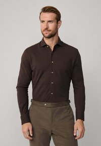 Hombre con cabello castaño claro que lleva una camisa de botones de manga larga marrón oscuro metida en pantalones verde oliva, de pie frente a un fondo liso.