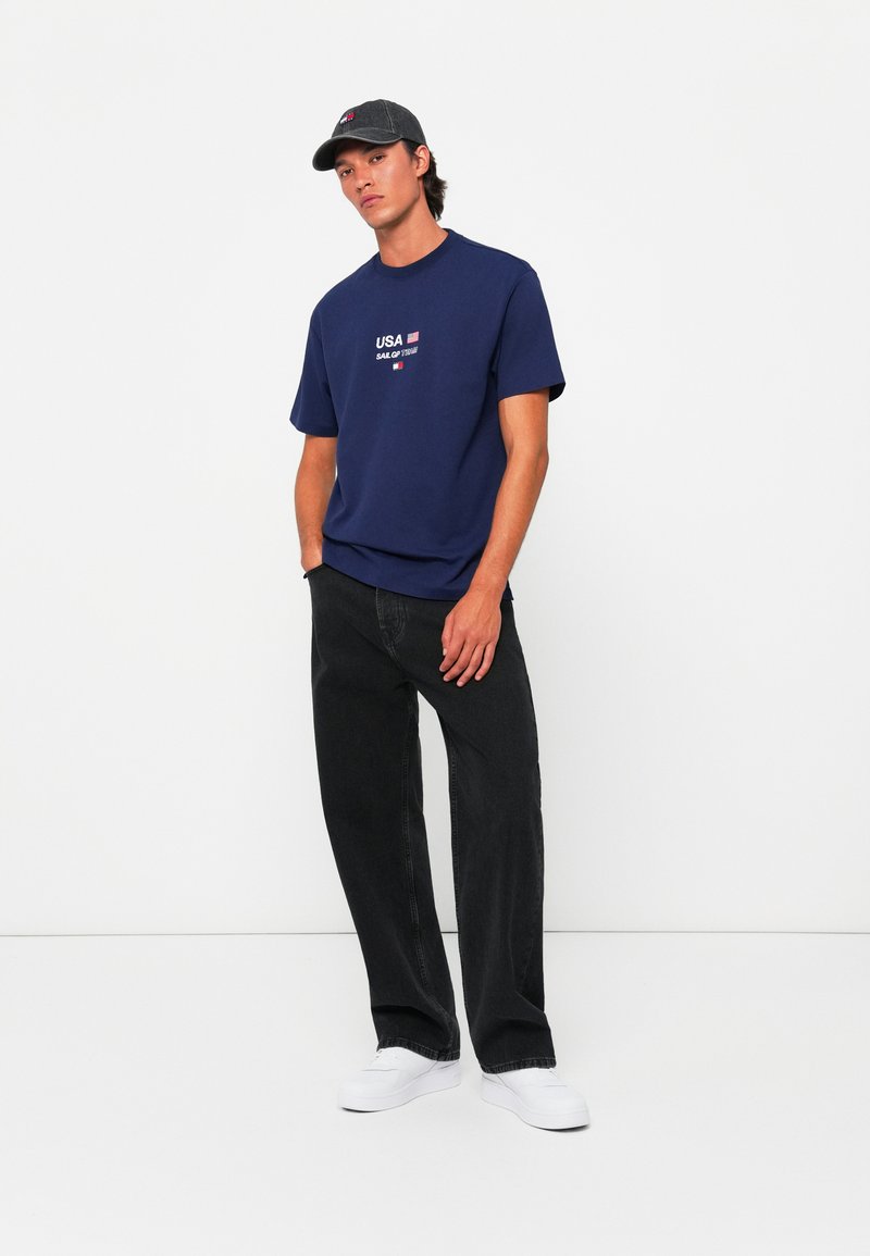 Navy blue T-shirt with white "USA" text and flag graphic, paired with black loose-fit jeans and white sneakers, accented by a gray cap.