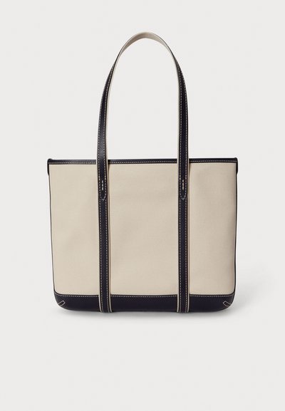 Plátenná taška na rameno v béžovej farbe s tmavomodrým koženým lemovaním, vybavená dvoma dlhými rúčkami, kontrastným prešívaním a pevnou obdĺžnikovou konštrukciou.