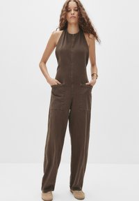 Donna con capelli ricci che indossa una tuta senza maniche marrone con ampie tasche anteriori e scarpe beige, in piedi su uno sfondo bianco semplice.