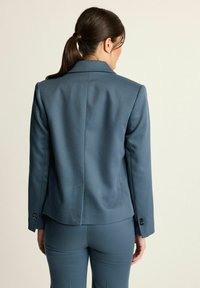 Vrouw met donker haar in een paardenstaart, gekleed in een getailleerde blauwe blazer en bijpassende broek, van zich af gekeerd tegen een lichte effen achtergrond.