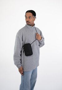 Gray fleece pullover with high collar, paired with light blue jeans. Black rectangular bag worn across the chest with adjustable strap.