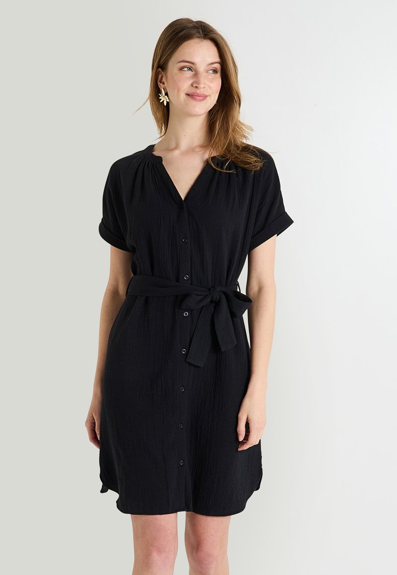 Vestido negro de manga corta con escote en V, cierre de botones y cintura anudada. Presenta un tejido texturizado y un diseño a la altura de la rodilla.
