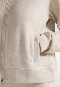 Beige zip-up hoodie med glittrande textur, med en framficka och ribbade ärmslut. Silverfärgad logotypdetalj nära fållen.