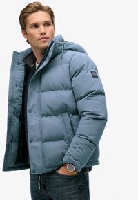 Lys blå pufferjakke med hætte, der har quiltede sektioner og trykknaplukninger. Bæres over et mørkt lag og parret med denim.