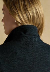 Tmavo sivý kabát s rybím vzorom a vysokým golierom, s diskrétnym logom vyšitou na chrbte, vyrobený z texturovanej vlnenej zmesi.