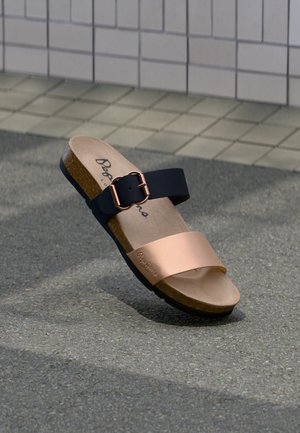 Basutė su kamštine pado dalimi, juoda reguliuojama dirželio sagtis ir metalinis rožinio aukso priekinis dirželis ant pilkos trinkelių fono.